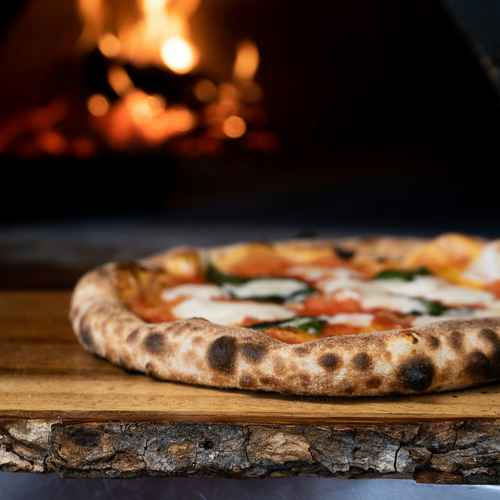 Eine gebackene Pizza auf einem Holzbrett vor einem Holzoffen mit Flamme